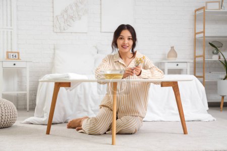 Young Asian woman preparing steam inhalation with herbs at table in bedroom