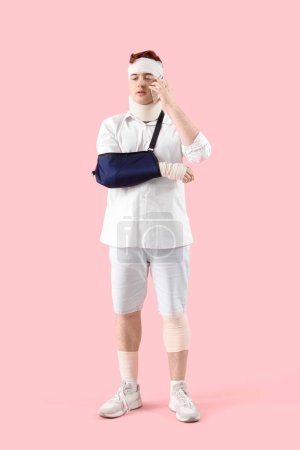 Injured young man after accident with broken arm talking by mobile phone on pink background