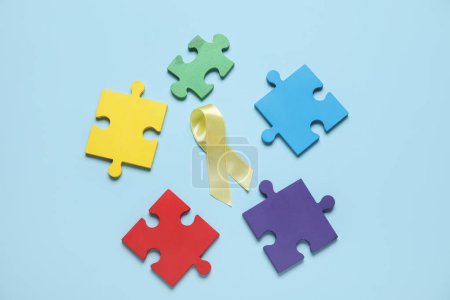 Téléchargez les photos : Ruban doré avec des pièces de puzzle sur fond bleu. Concept de sensibilisation au cancer chez l'enfant - en image libre de droit
