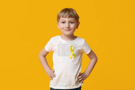 Téléchargez les photos : Mignon petit garçon avec ruban jaune sur fond de couleur. Journée internationale du cancer chez les enfants - en image libre de droit