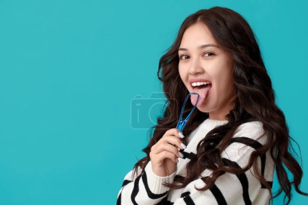 Beautiful young Asian woman with tongue scraper on blue background
