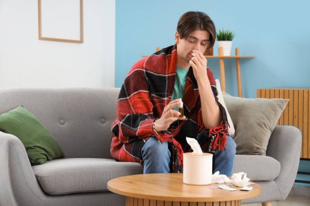Téléchargez les photos : Jeune homme malade avec des gouttes nasales assis sur le canapé à la maison - en image libre de droit