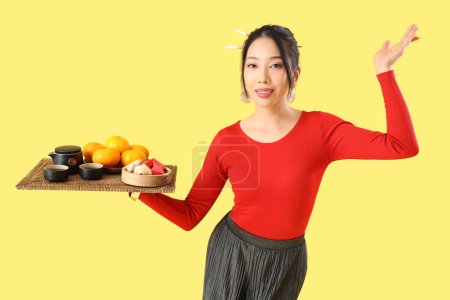 Photo for Young Asian woman holding tray with tea, mandarins and fortune cookies on yellow background. Chinese New Year celebration - Royalty Free Image