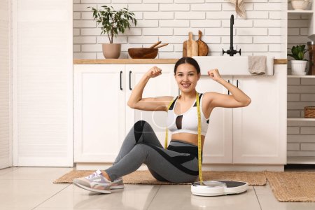 Téléchargez les photos : Jeune femme en vêtements de sport et avec ruban à mesurer assis près des balances dans la cuisine. Concept de perte de poids - en image libre de droit