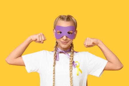 Téléchargez les photos : Adolescente mignonne en costume de super-héros avec ruban jaune sur fond de couleur. Journée internationale du cancer chez les enfants - en image libre de droit