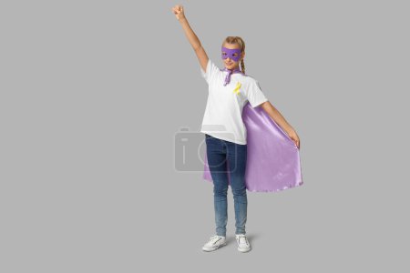 Téléchargez les photos : Adolescente mignonne en costume de super-héros avec ruban jaune sur fond gris. Journée internationale du cancer chez les enfants - en image libre de droit