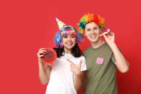 Young friends in funny disguise with party whistle pointing at whoopee cushion on red background. April fool's day celebration