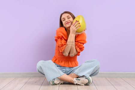 Foto de Mujer joven sonriente con pomelo sentado cerca de la pared lila - Imagen libre de derechos