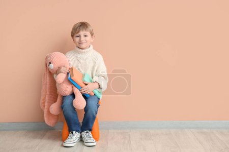 Foto de Lindo niño pequeño con juguete conejito y libros sentados en silla contra la pared de color - Imagen libre de derechos