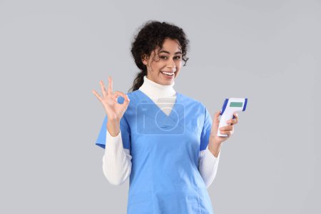 Pasante médica afroamericana femenina con termómetro infrarrojo que muestra OK sobre fondo claro