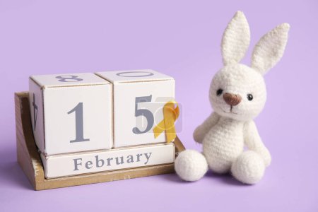 Téléchargez les photos : Calendrier avec la date de la journée de sensibilisation au cancer de l'enfance, ruban doré et lapin jouet sur fond lilas - en image libre de droit