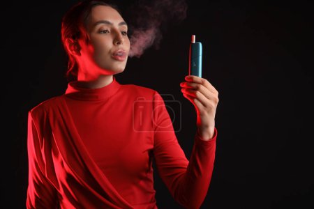 Téléchargez les photos : Belle jeune femme fumant cigare électronique sur fond sombre - en image libre de droit