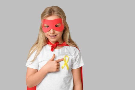 Téléchargez les photos : Adolescente mignonne en costume de super-héros pointant vers le ruban jaune sur fond gris. Journée internationale du cancer chez les enfants - en image libre de droit