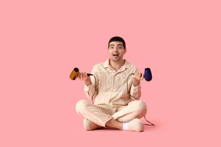 Handsome young man with hair dryers sitting against pink background