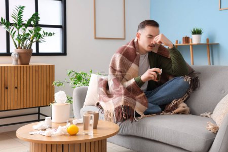 Téléchargez les photos : Jeune homme malade avec plaid et gouttes nasales assis sur le canapé à la maison - en image libre de droit
