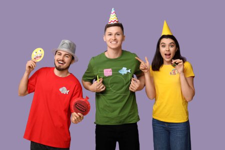 Young friends in funny disguise with paper fishes and whoopee cushion on purple background. April fool's day celebration