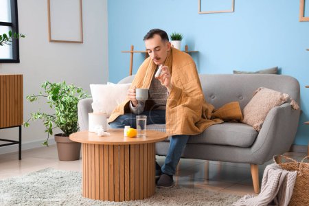 Téléchargez les photos : Jeune homme malade avec une tasse de thé et des gouttes nasales assis sur le canapé à la maison - en image libre de droit