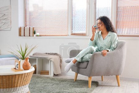 Young African-American woman using asthma inhaler in in armchair at home