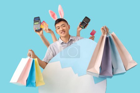 Junger asiatischer Mann in Hasenohren mit Einkaufstaschen und Papierei auf blauem Hintergrund. Osterverkauf