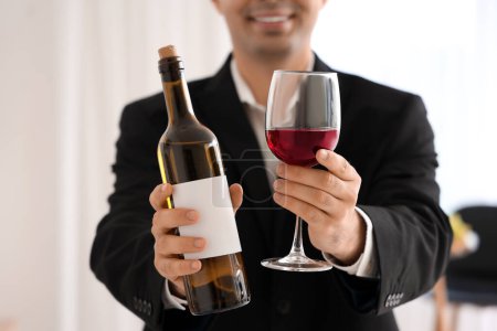 Photo for Young sommelier with bottle and glass of red wine in kitchen, closeup - Royalty Free Image