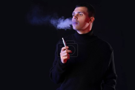 Téléchargez les photos : Jeune homme fumant cigarette électronique sur fond noir - en image libre de droit