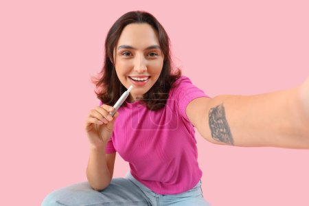 Young woman with electronic cigar taking selfie on pink background, closeup