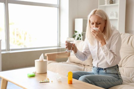 Sick mature woman with tissues, pills and glass of water at home