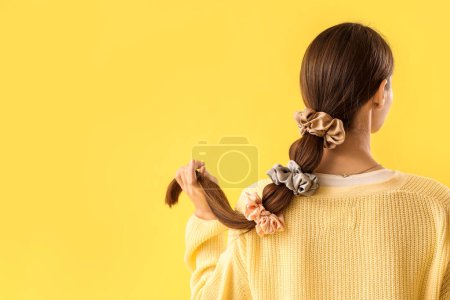 Beautiful young woman with silk scrunchies on ponytail against yellow background