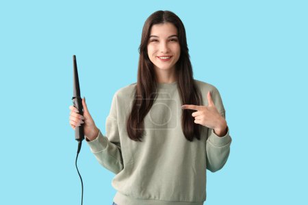 Beautiful young happy woman pointing at curling iron on blue background