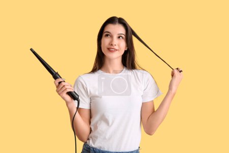 Beautiful young happy woman with curling iron on yellow background