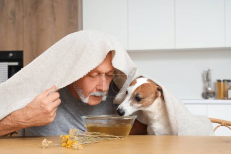 Mature man with dog doing steam inhalation at table in kitchen