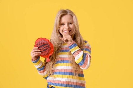 Funny little girl with whoopee cushion showing silence gesture on yellow background. April Fools' Day celebration