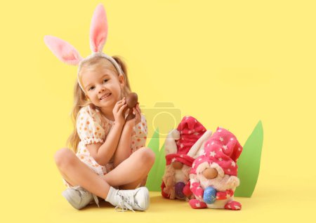 Photo for Cute little girl in bunny ears with chocolate Easter egg and gnomes sitting on yellow background - Royalty Free Image