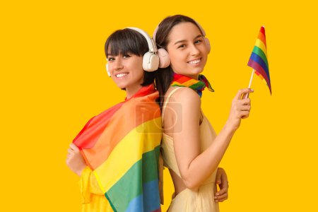 Foto de Pareja joven lesbiana en auriculares con banderas LGBT sobre fondo amarillo - Imagen libre de derechos