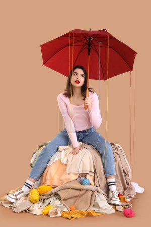 Young woman with umbrella and knitting yarn sitting on heap of clothes against beige background