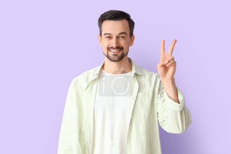 Téléchargez les photos : Bel homme montrant geste de victoire sur fond lilas - en image libre de droit
