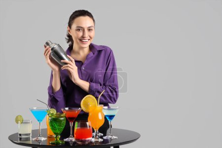 Female bartender with shaker and cocktails at table on light background