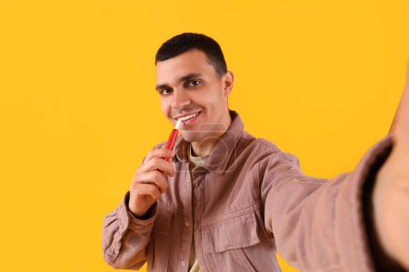 Téléchargez les photos : Jeune homme avec prise de cigarette électronique selfie sur fond jaune - en image libre de droit