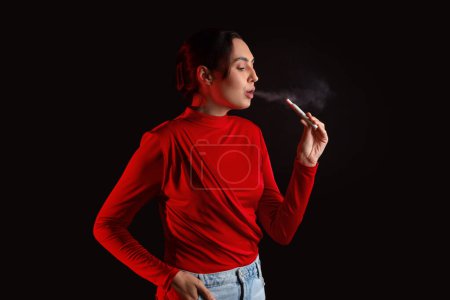 Téléchargez les photos : Belle jeune femme fumant cigare électronique sur fond sombre - en image libre de droit
