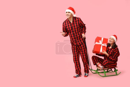 Happy young couple in Christmas pajamas, with sledge and gift on pink background