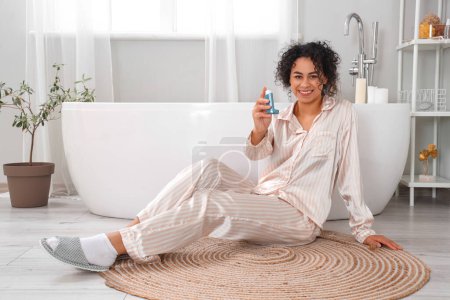 Téléchargez les photos : Sourire jeune femme afro-américaine avec inhalateur d'asthme assis sur le sol dans la salle de bain - en image libre de droit