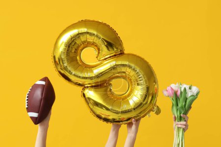 Female hands with foil balloon in shape of figure 8, rugby ball and tulip flowers for International Women's Day on yellow background