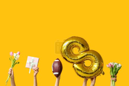 Female hands with foil balloon in shape of figure 8, rugby ball and tulip flowers on yellow background. International Women's Day