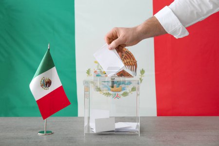 Junger Mann mit mexikanischer Flagge in der Nähe der Wahlurne auf einem Tisch im Wahllokal, Nahaufnahme