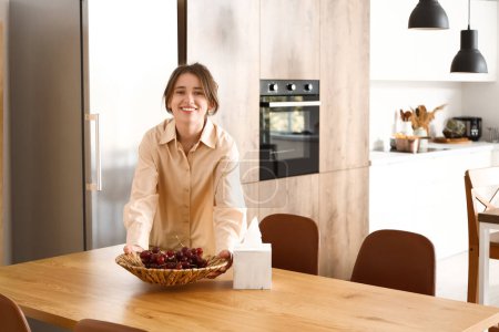 Photo for Pretty young woman with bowl of grapes on wooden table in modern kitchen - Royalty Free Image