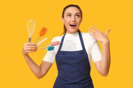 Portrait of happy young housewife in apron with kitchen utensils on yellow background