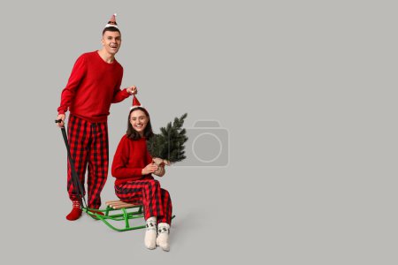 Happy young couple in Christmas pajamas, with sledge and coniferous tree on grey background