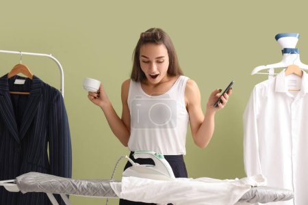 Shocked young businesswoman with smartphone and cup of coffee noticed iron on ironing board at home