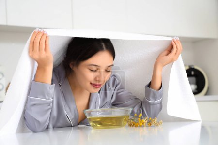 Young Asian woman doing steam inhalation at table in kitchen