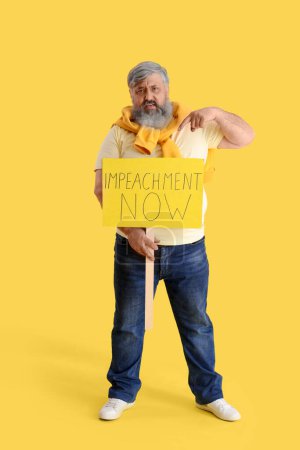 Protesting mature man pointing at placard with text IMPEACHMENT NOW on yellow background